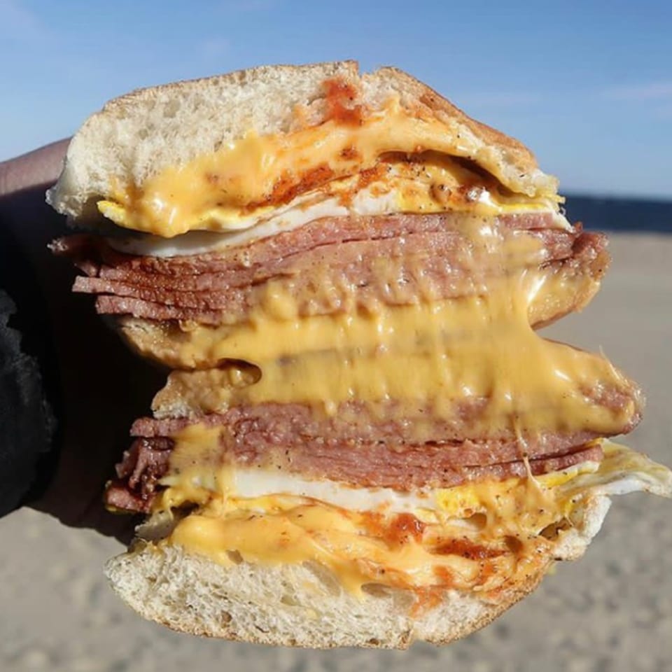 A sliced Bing's Beach House Deli sandwich with meat, eggs, and melted cheese, held outdoors.